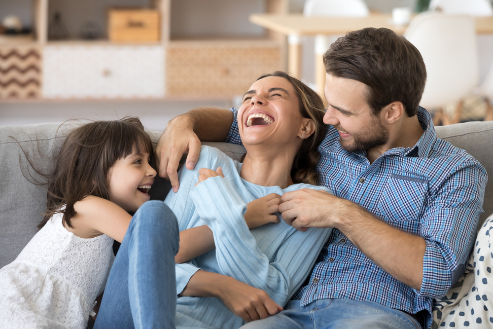 A family shares a joyful laugh on the couch, their smiles radiating happiness and good dental health, thanks to Ates Dental Studio
